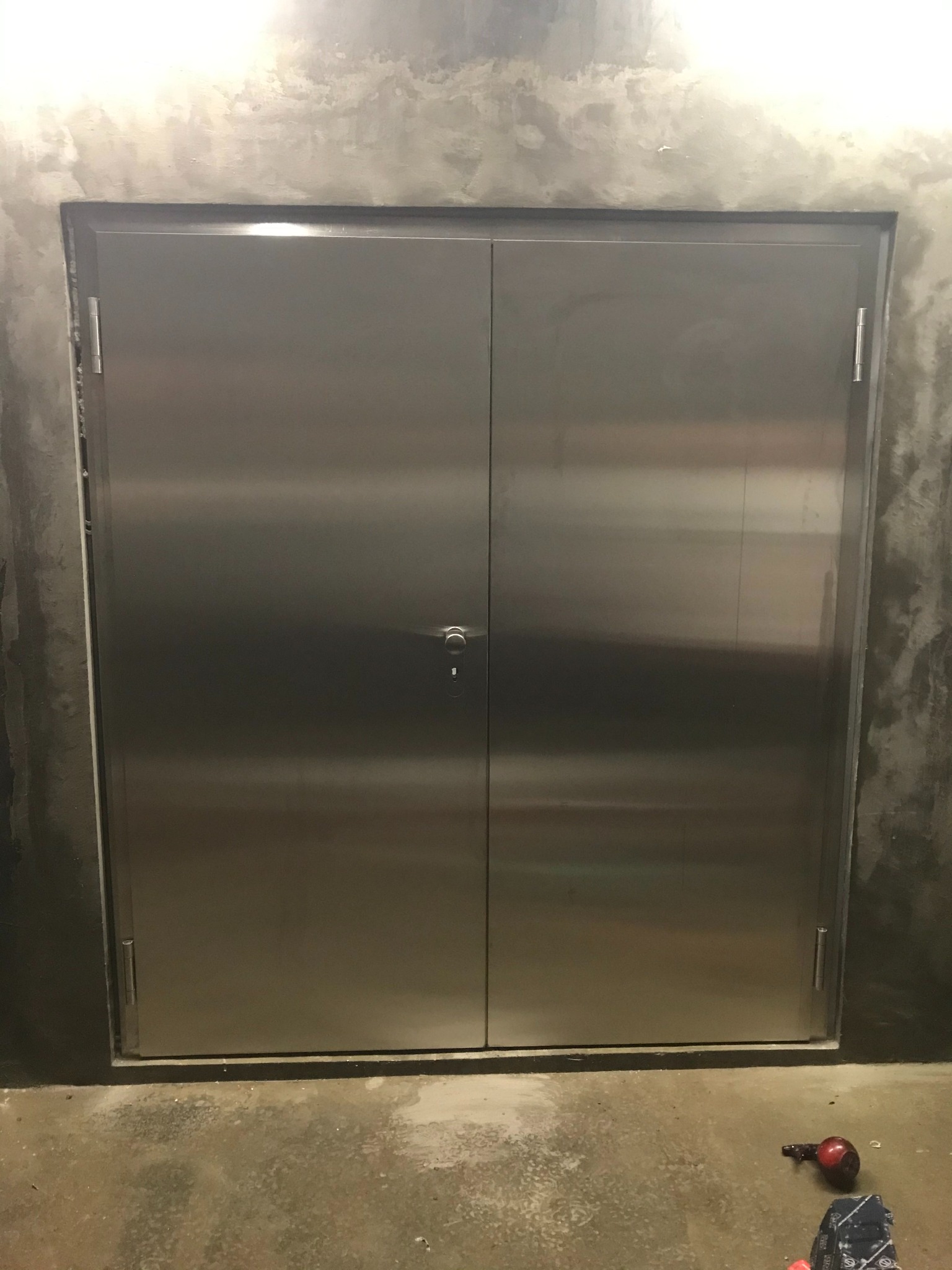 Metalowe, dwuskrzydłowe drzwi w surowym, betonowym otoczeniu. Minimalistyczny design, widoczne zawiasy i klamka. Podłoga betonowa z drobnymi zabrudzeniami.