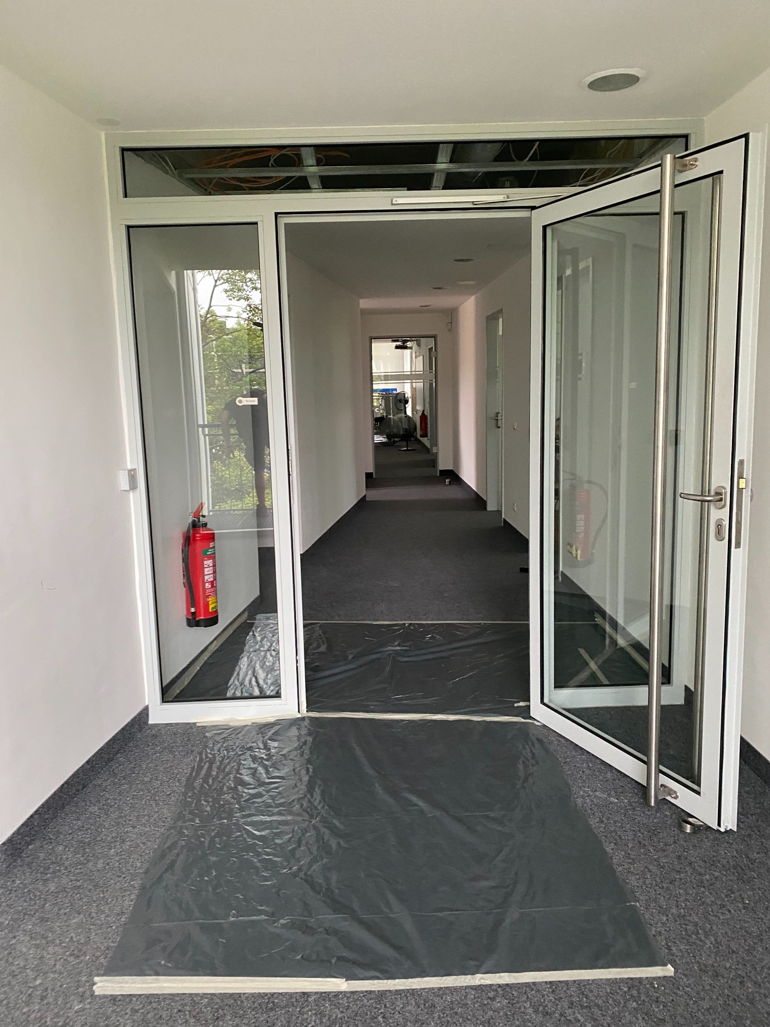 Białe, przeszklone drzwi wejściowe do biura, częściowo otwarte, z widoczną instalacją nadprożową i zabezpieczeniem podłogi folią malarską. Widok na korytarz.