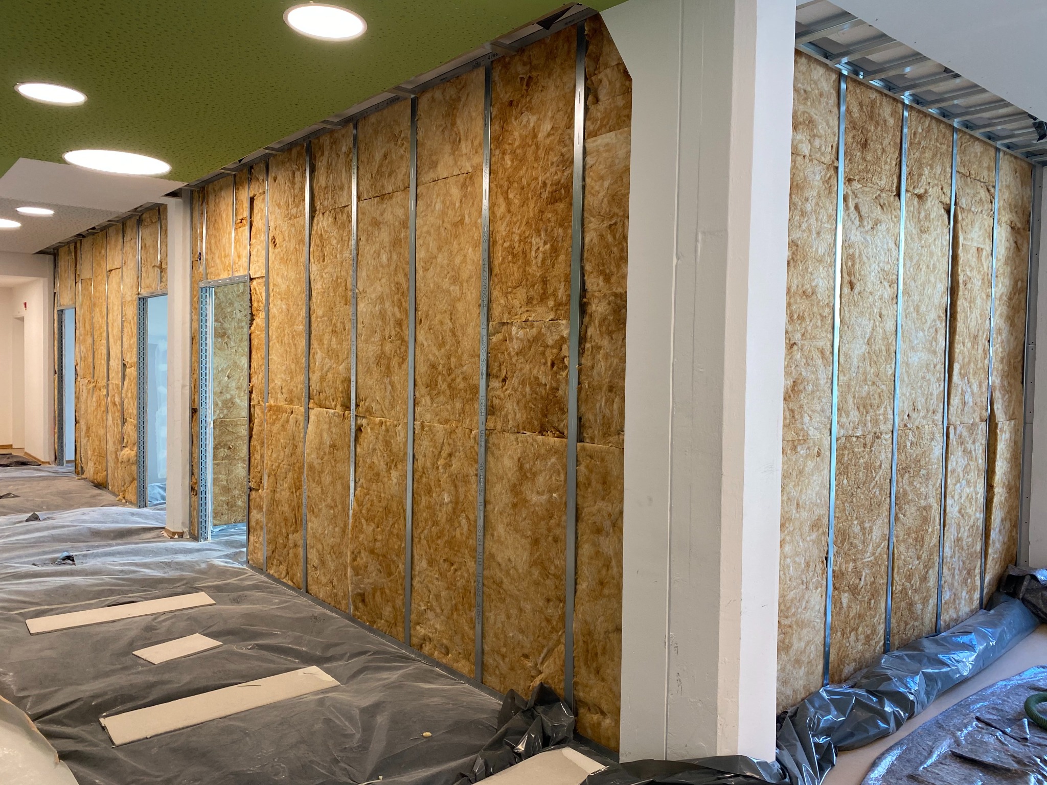 Ściana z izolacją z wełny mineralnej w metalowym stelażu, widoczne profile i fragment sufitu z oświetleniem. Podłoga zabezpieczona folią malarską.