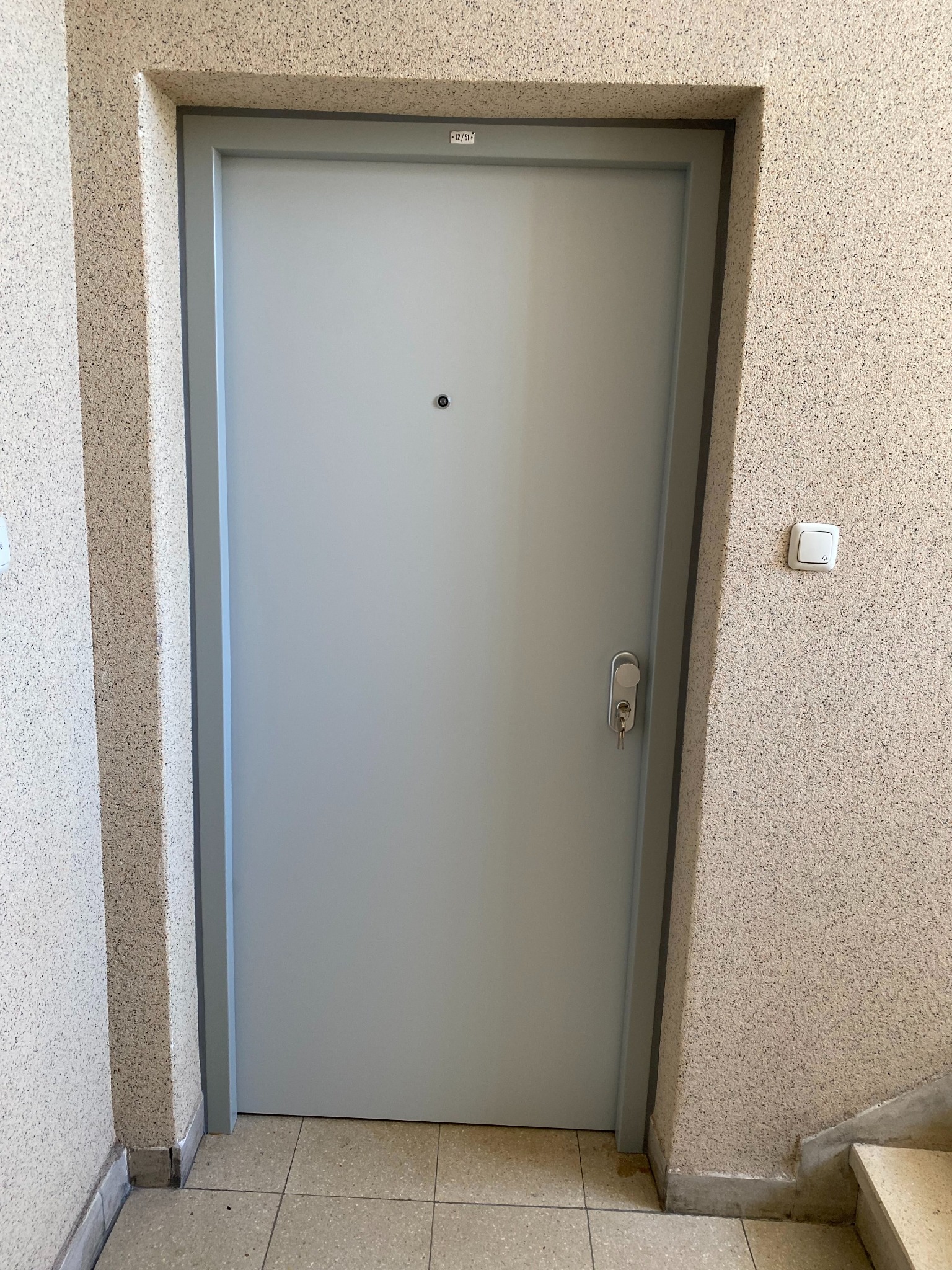 Szare, metalowe drzwi wejściowe z wizjerem i kluczem w zamku, osadzone w ścianie o fakturze baranka. Widoczny fragment klatki schodowej i włącznik dzwonka.