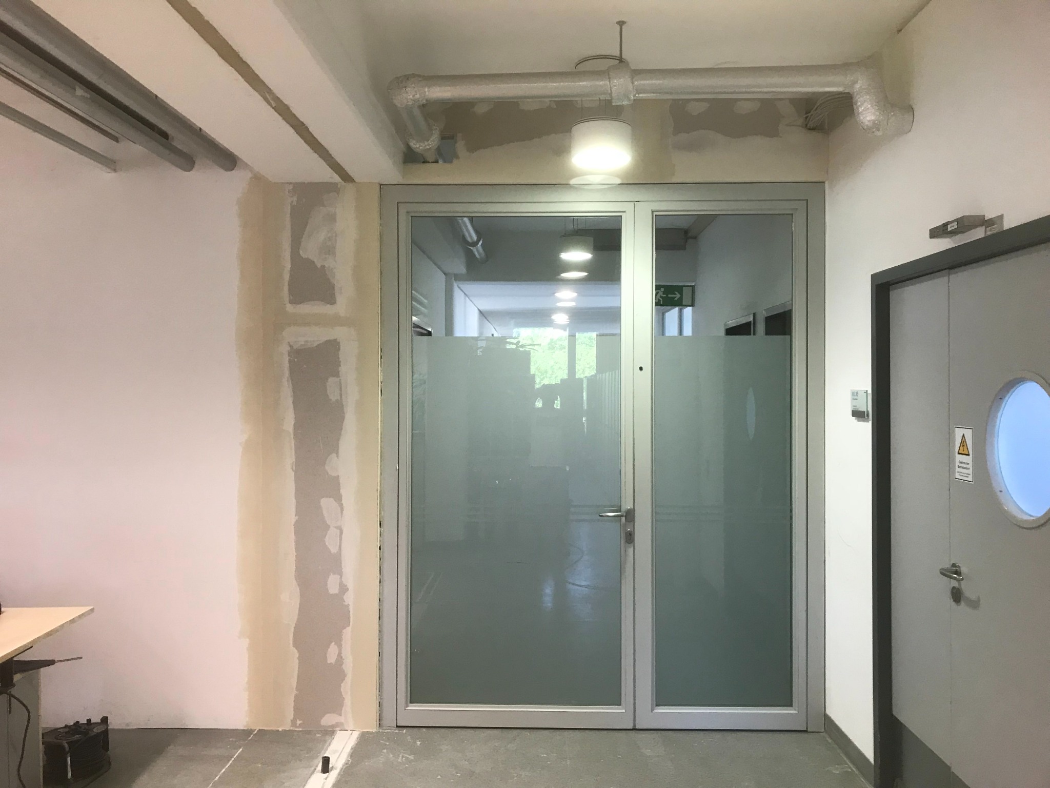 Korytarz w trakcie prac wykończeniowych: ściana z widocznymi śladami szpachlowania, aluminiowe drzwi z matową szybą, instalacja pod sufitem.