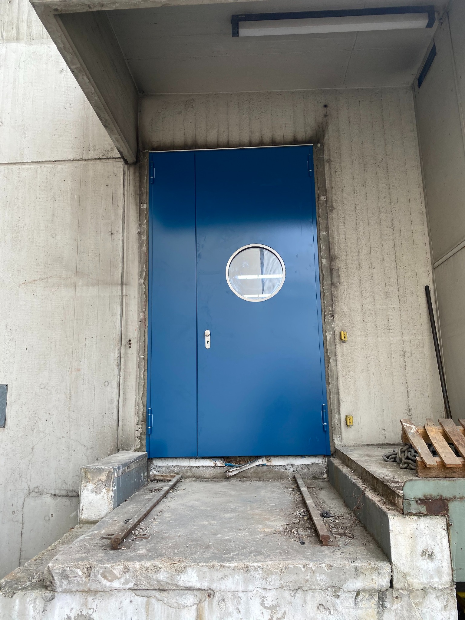 Niebieskie, metalowe drzwi z okrągłym okienkiem w surowym, betonowym otoczeniu. Widoczne ślady użytkowania i elementy konstrukcyjne.