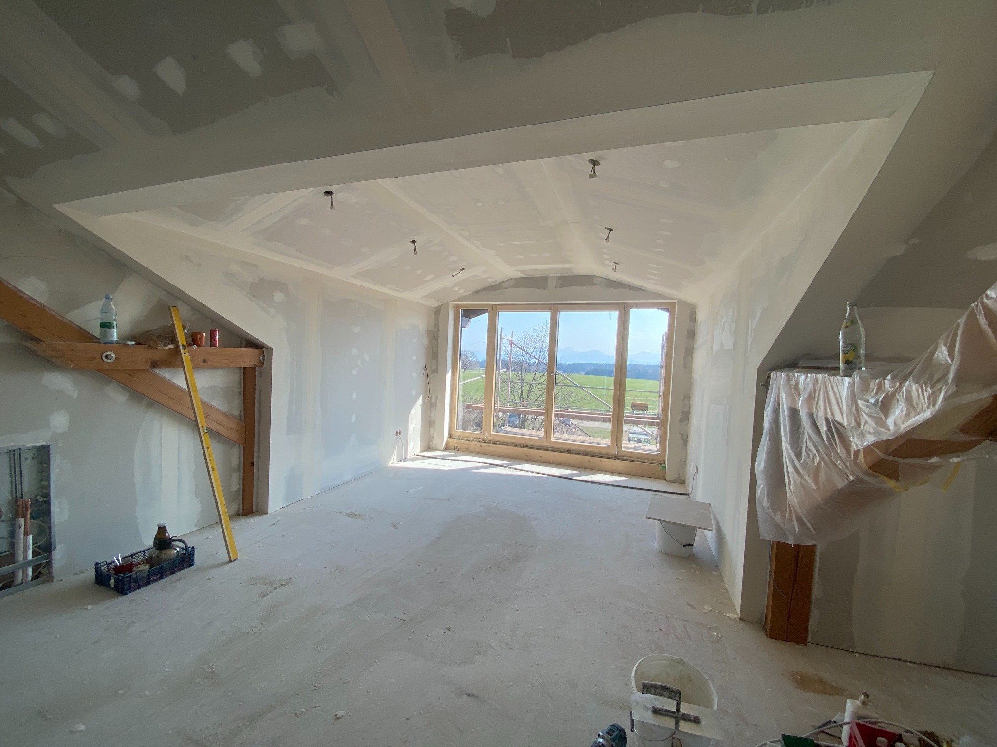 Poddasze po szpachlowaniu i montażu płyt gipsowych, z oknem na krajobraz. Widoczne narzędzia i materiały budowlane, surowy stan wykończenia.