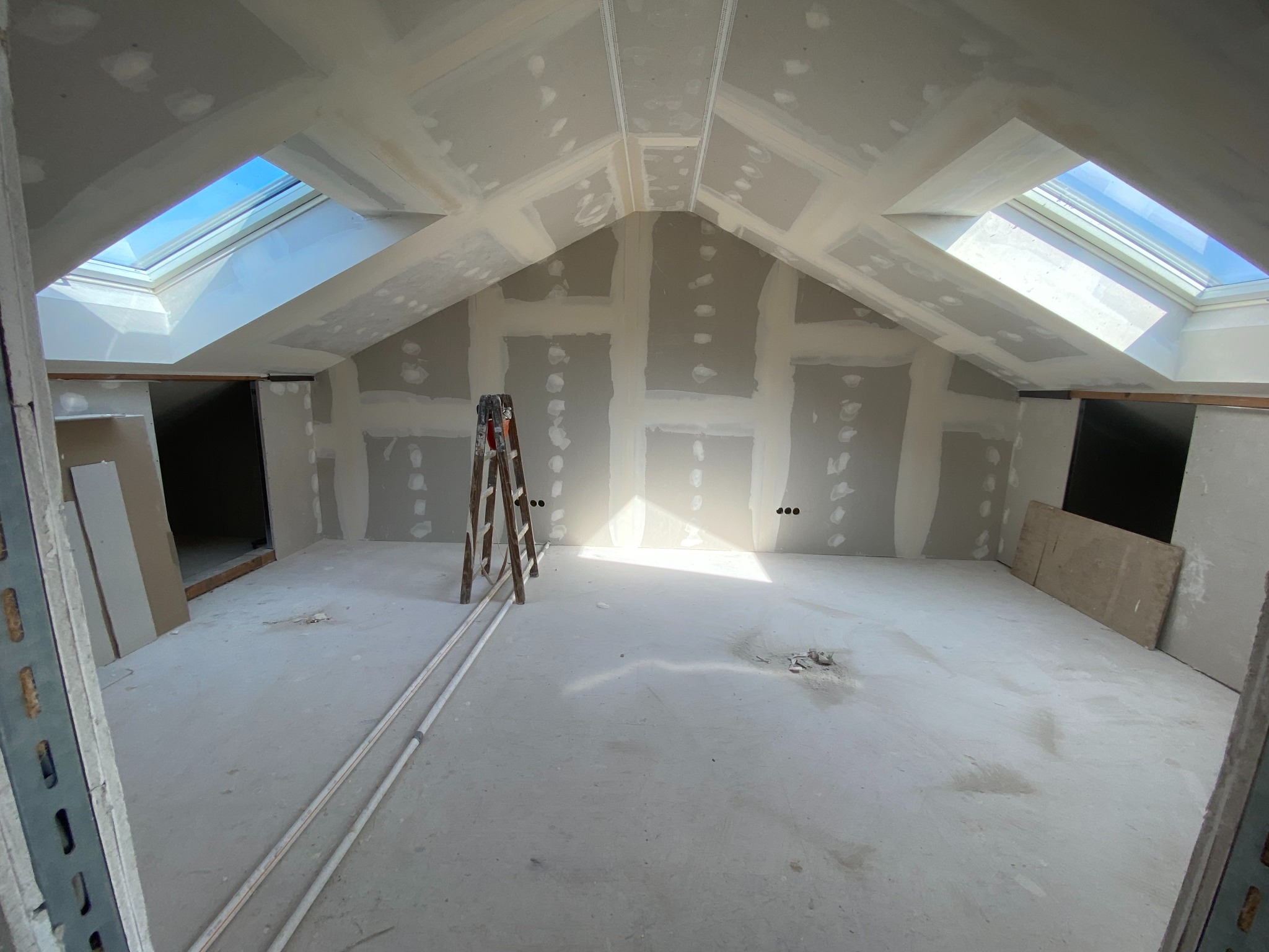 Poddasze w trakcie wykańczania: ściany i sufit pokryte płytami gipsowo-kartonowymi, widoczne łączenia szpachlowane, drabina w centrum, okna dachowe w skosach.