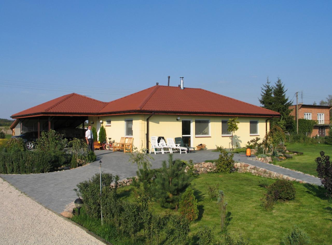 Parterowy dom z czerwonym dachem i żółtą elewacją, widok z ogrodu. Na podjeździe stoi samochód, obok domu drewniane meble ogrodowe i leżaki.