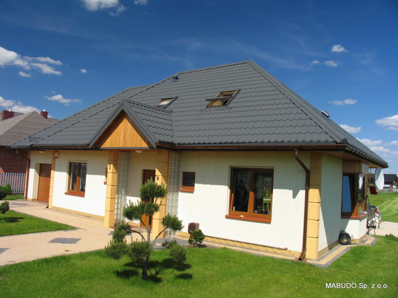 Gotowy dom jednorodzinny z szarą dachówką i jasną elewacją, widok z zewnątrz w słoneczny dzień. Elementy dekoracyjne z drewna i szkła.