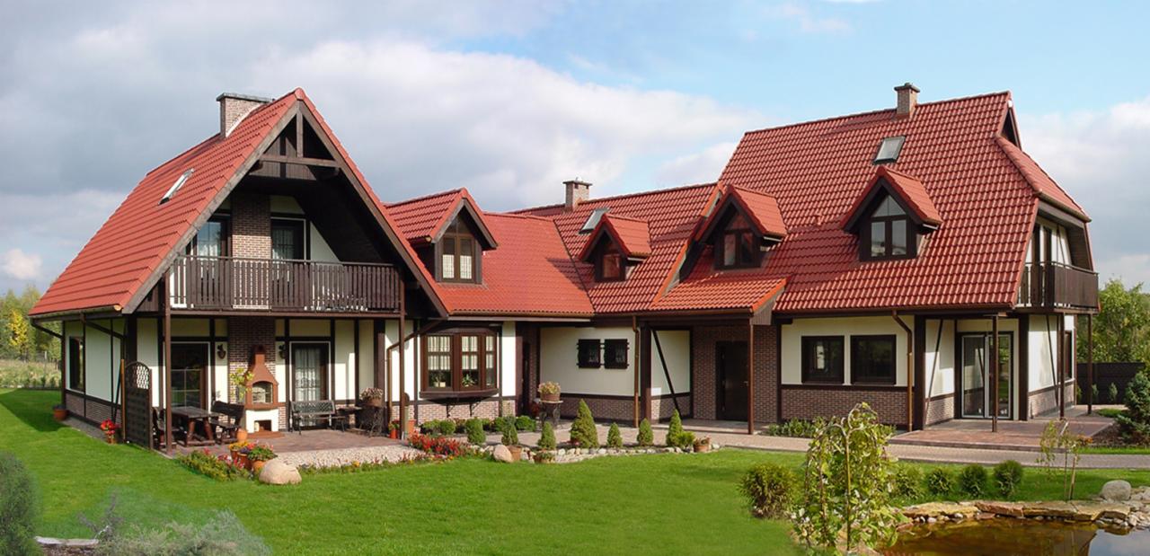 Szeroki kadr na dom szkieletowy z czerwoną dachówką, widoczny z zadbanego ogrodu z oczkiem wodnym i kwiatami, w słoneczny dzień.