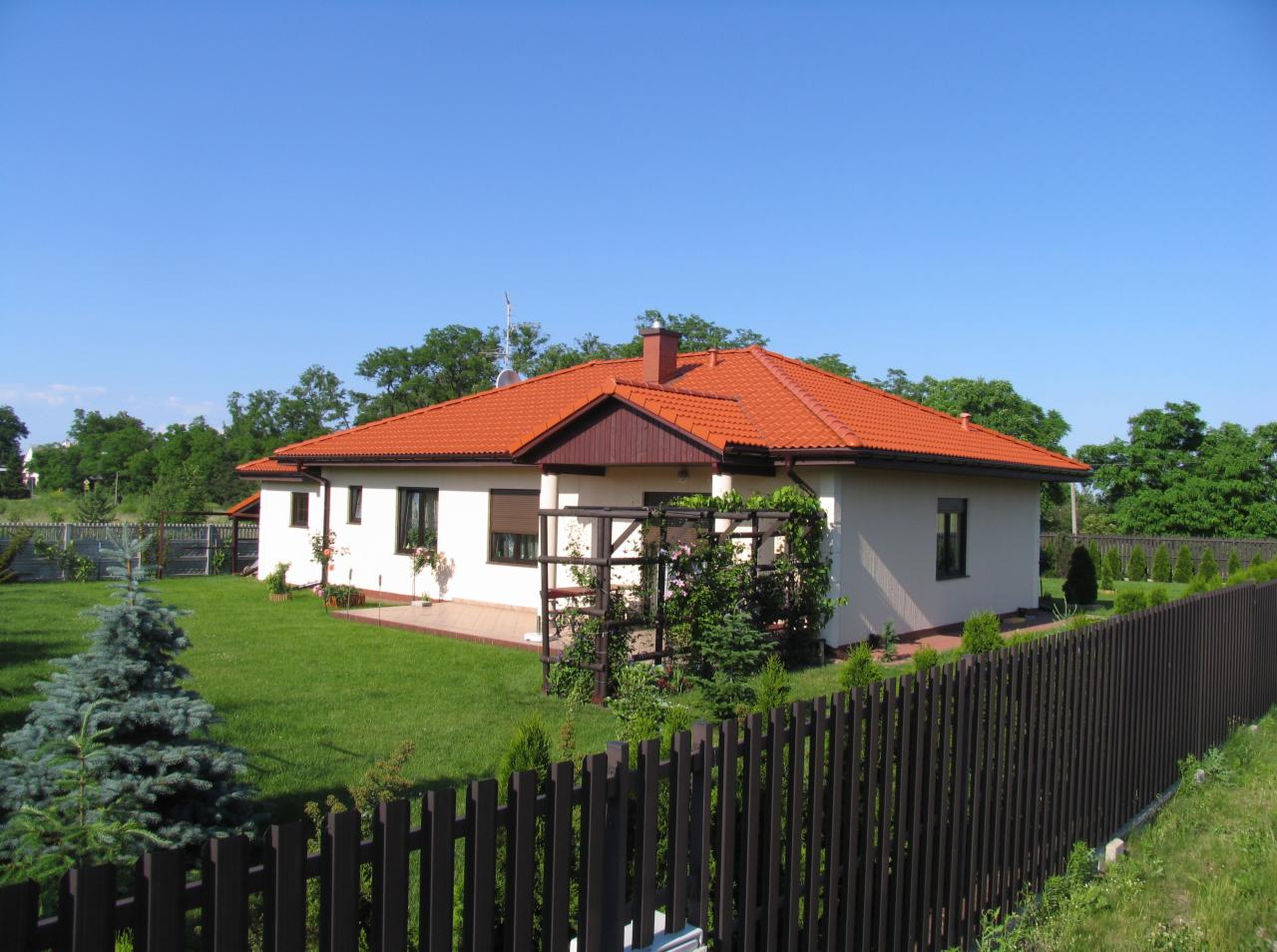 Parterowy dom z czerwoną dachówką, drewnianym ogrodzeniem i zadbanym trawnikiem w słoneczny dzień. Widoczne elementy architektury ogrodowej.