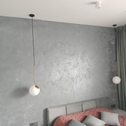 WYKOŃCZENIA WNĘTRZ PAWEŁ DZIURA - Ściana w pokoju sypialnym wykończona dekoracyjnym tynkiem w odcieniach szarości, oświetlona wiszącymi lampami z kulistymi kloszami i złotymi elementami, widoczny fragment łóżka z szarym zagłówkiem...
