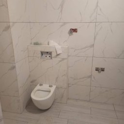 WYKOŃCZENIA WNĘTRZ PAWEŁ DZIURA - Świeżo wykafelkowana łazienka z podwieszaną toaletą, widoczne niedokończone podłączenia baterii i papier toaletowy na zabudowie stelaża.