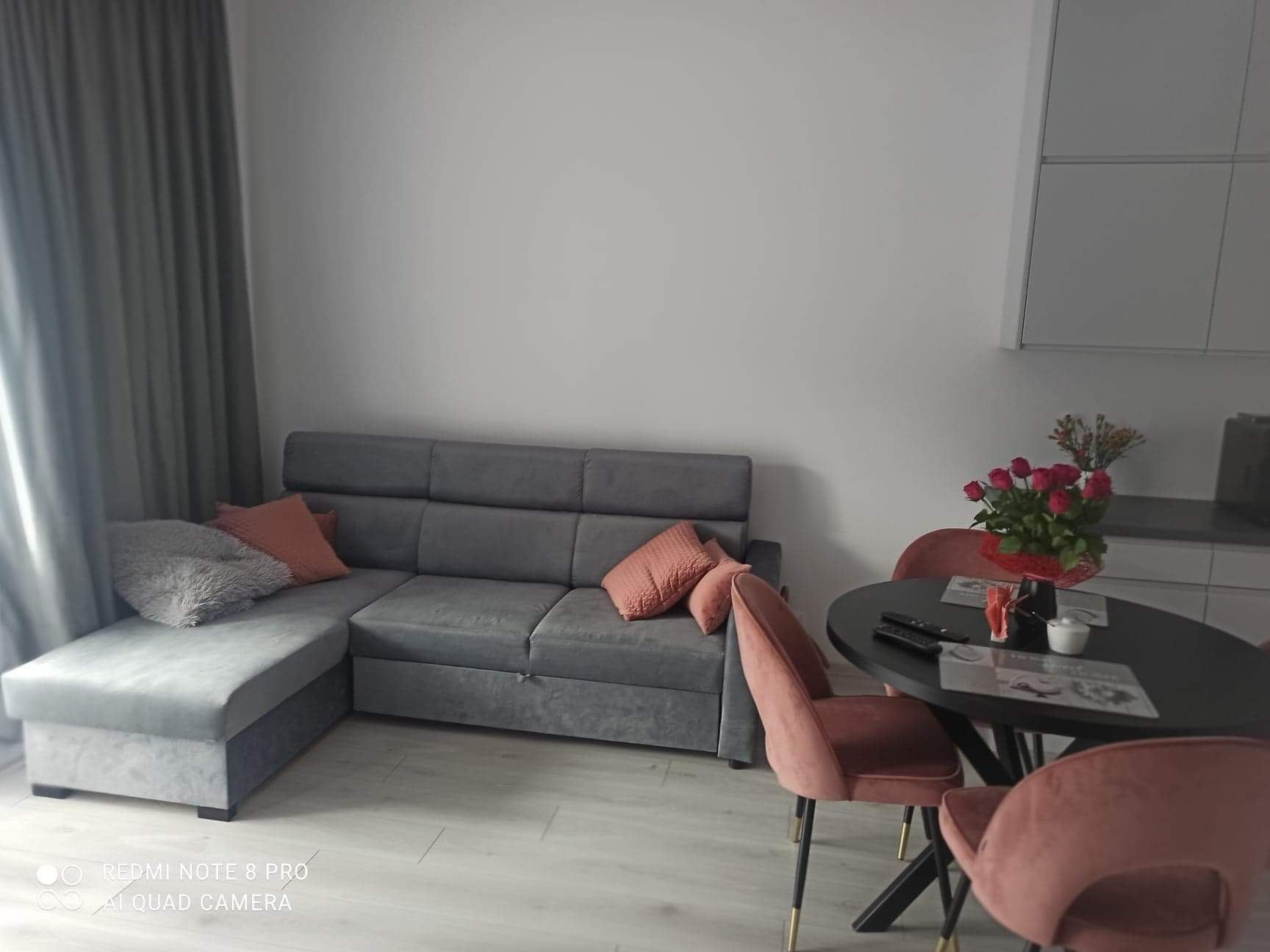 Szary narożnik z poduszkami w odcieniach różu i szarości w salonie z okrągłym stołem, różowymi krzesłami i bukietem czerwonych róż.