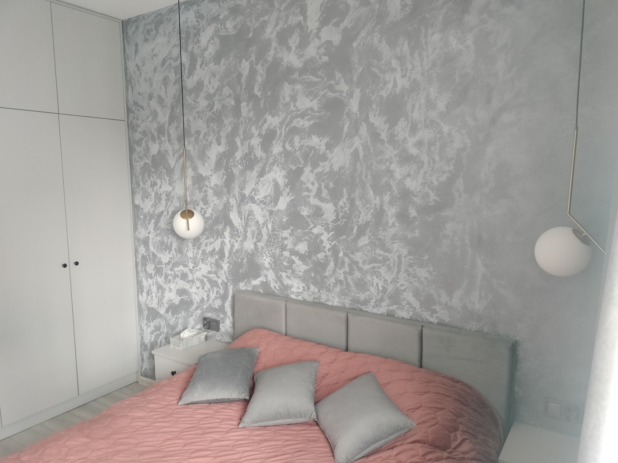 Sypialnia z łóżkiem w odcieniach różu i szarości, ściana z dekoracyjnym malowaniem w odcieniach srebra, wiszące lampy kuliste i biała szafa w zabudowie.