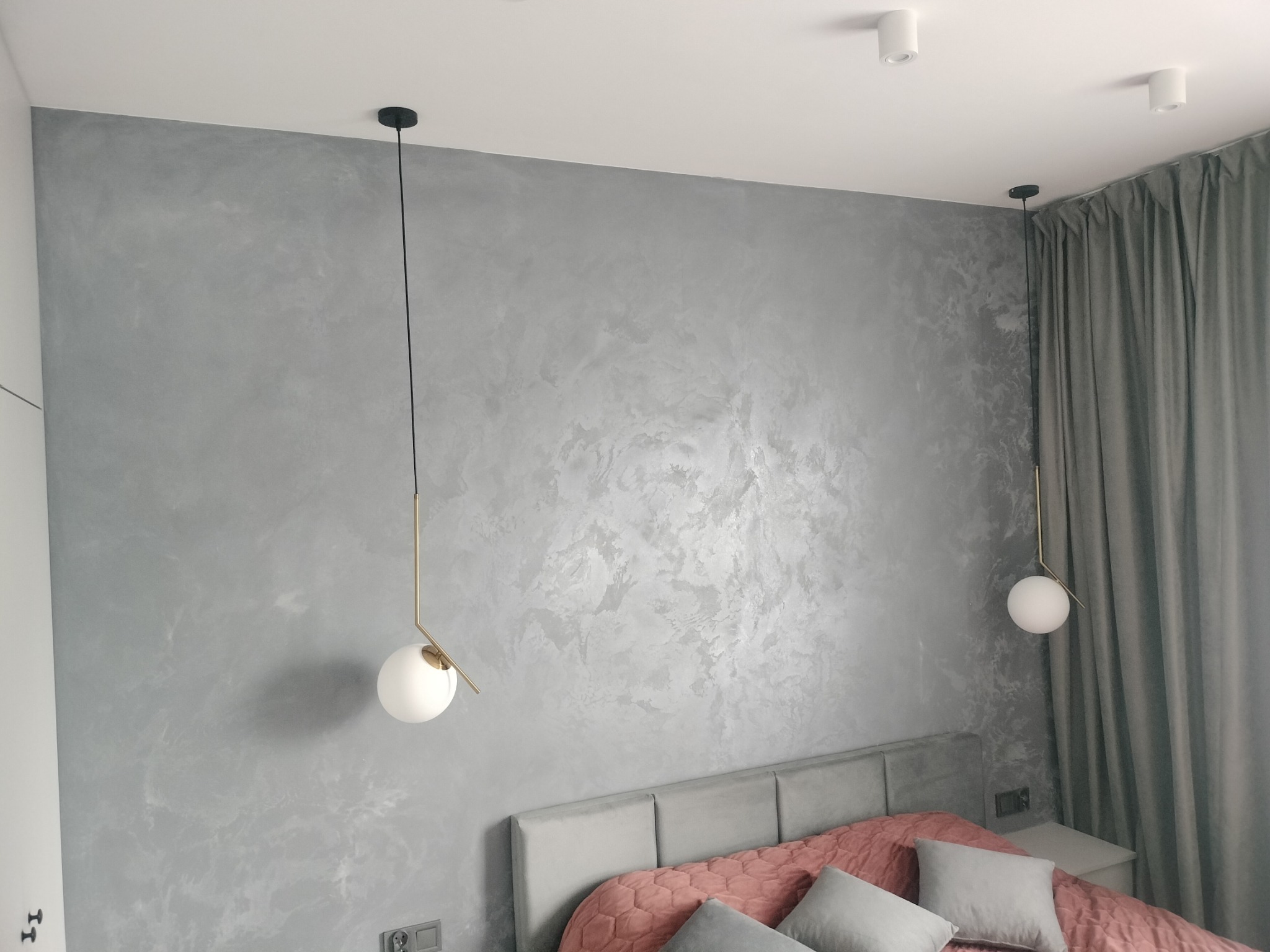 Ściana w pokoju sypialnym wykończona dekoracyjnym tynkiem w odcieniach szarości, oświetlona wiszącymi lampami z kulistymi kloszami i złotymi elementami, widoczny fragment łóżka z szarym zagłówkiem...