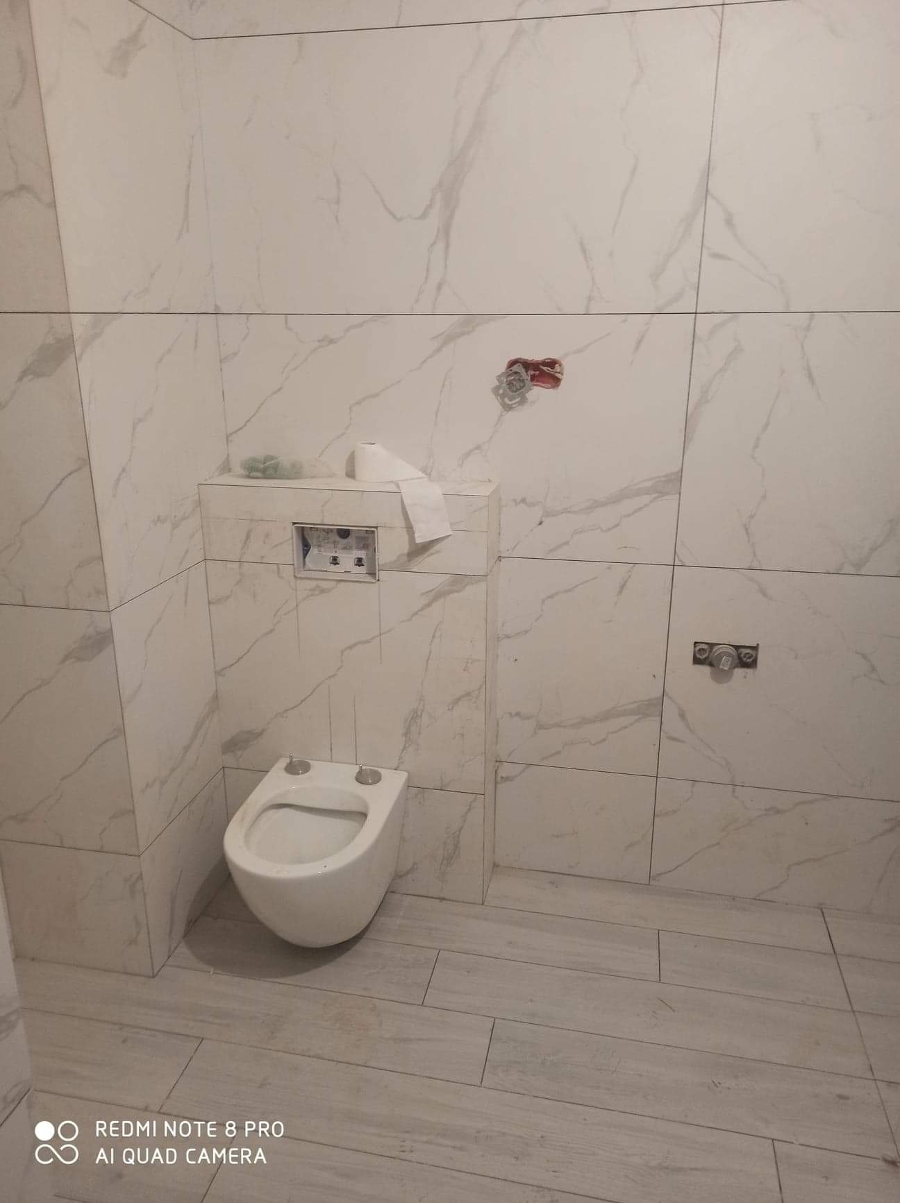 Świeżo wykafelkowana łazienka z podwieszaną toaletą, widoczne niedokończone podłączenia baterii i papier toaletowy na zabudowie stelaża.