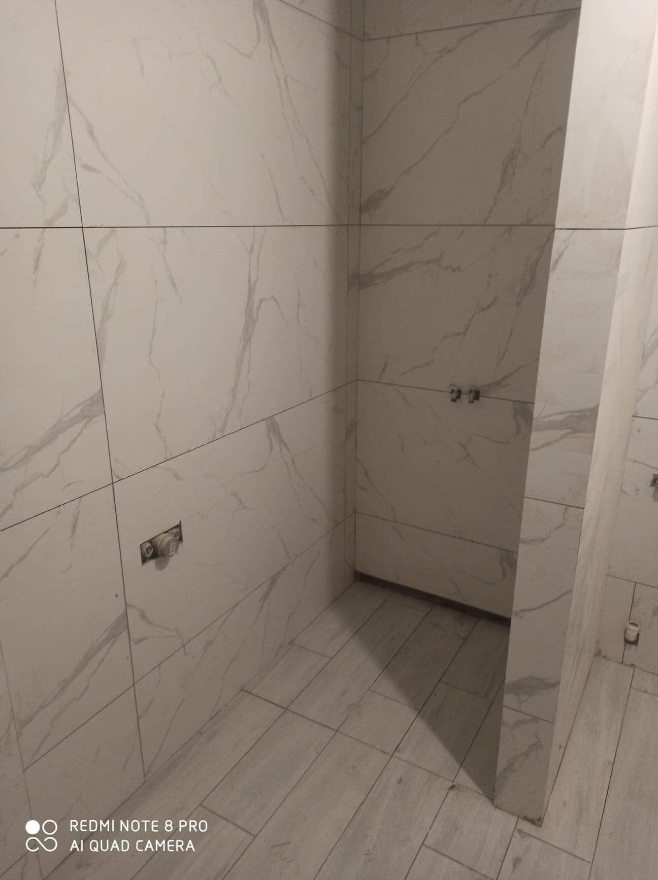 Wnętrze łazienki w trakcie wykańczania w Jarczowie, z położonymi białymi płytkami imitującymi marmur na ścianach i podłodze, widoczne wystające rury instalacji wodnej.