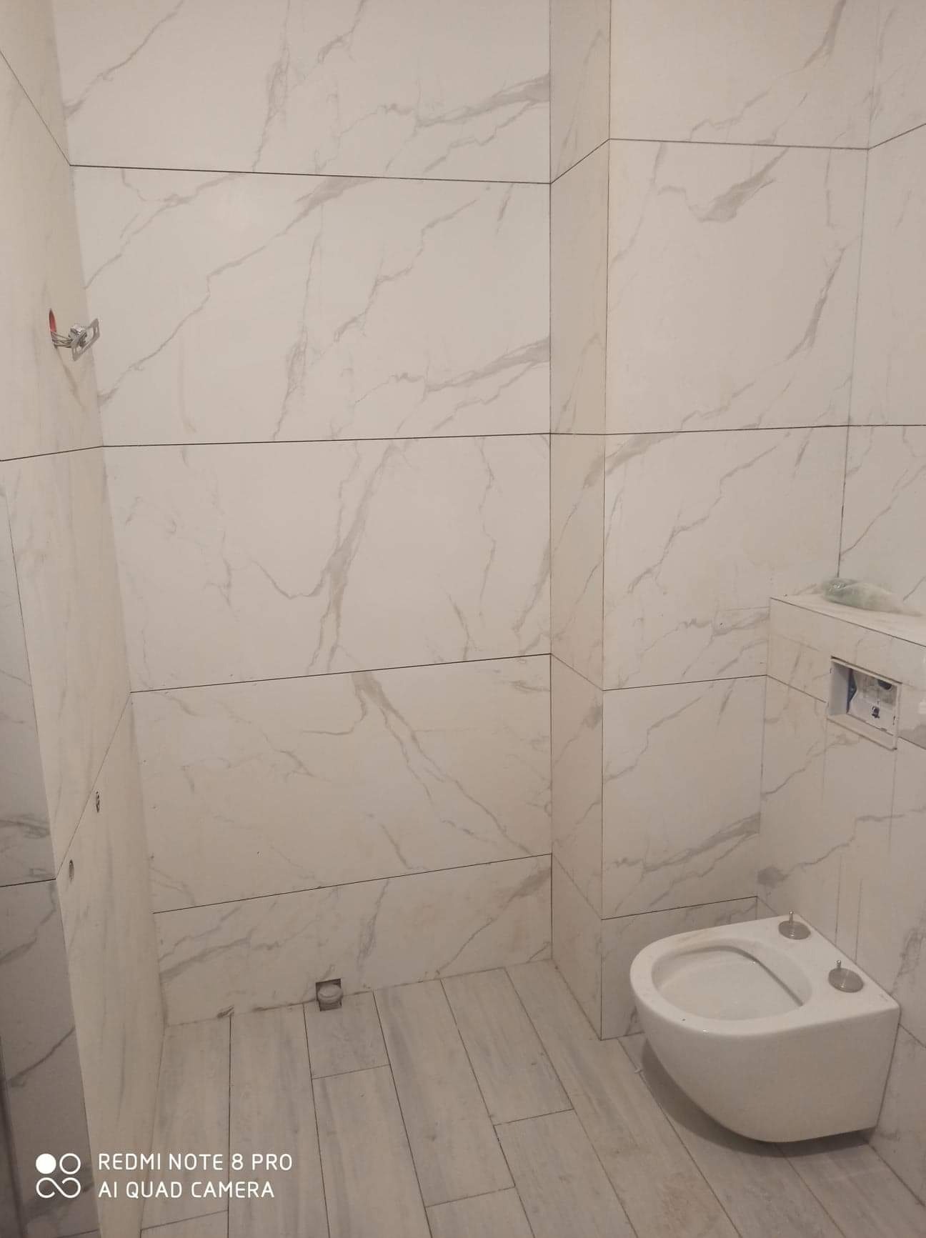 Świeżo wykafelkowana łazienka w odcieniach bieli z marmurkowym wzorem, z wiszącą miską WC i widocznymi elementami instalacji.