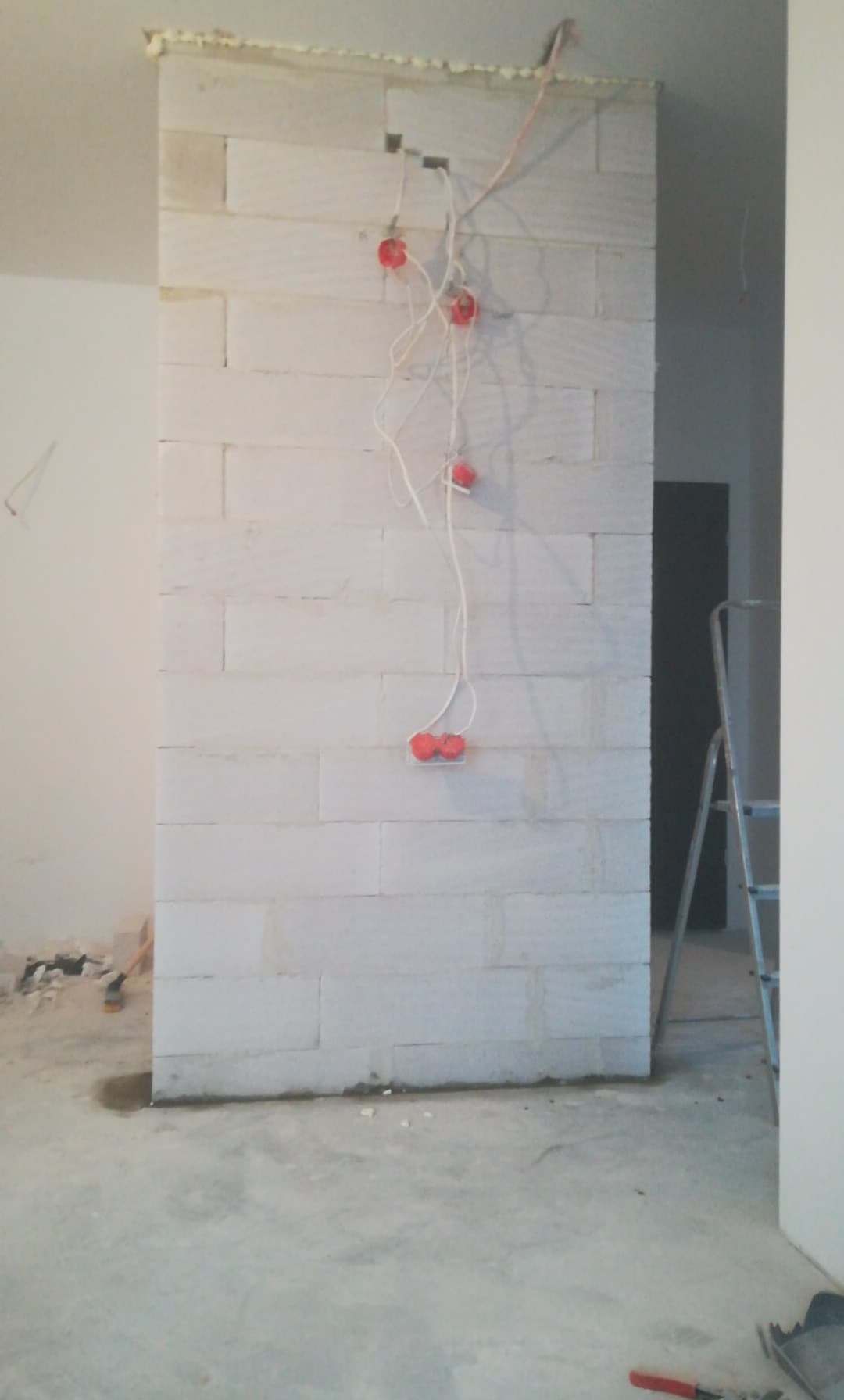 Wznoszona ściana działowa z bloczków z betonu komórkowego, widoczne puszki elektryczne z wystającymi przewodami, pianka montażowa na górze ściany, drabina w tle, surowy stan wykończenia.