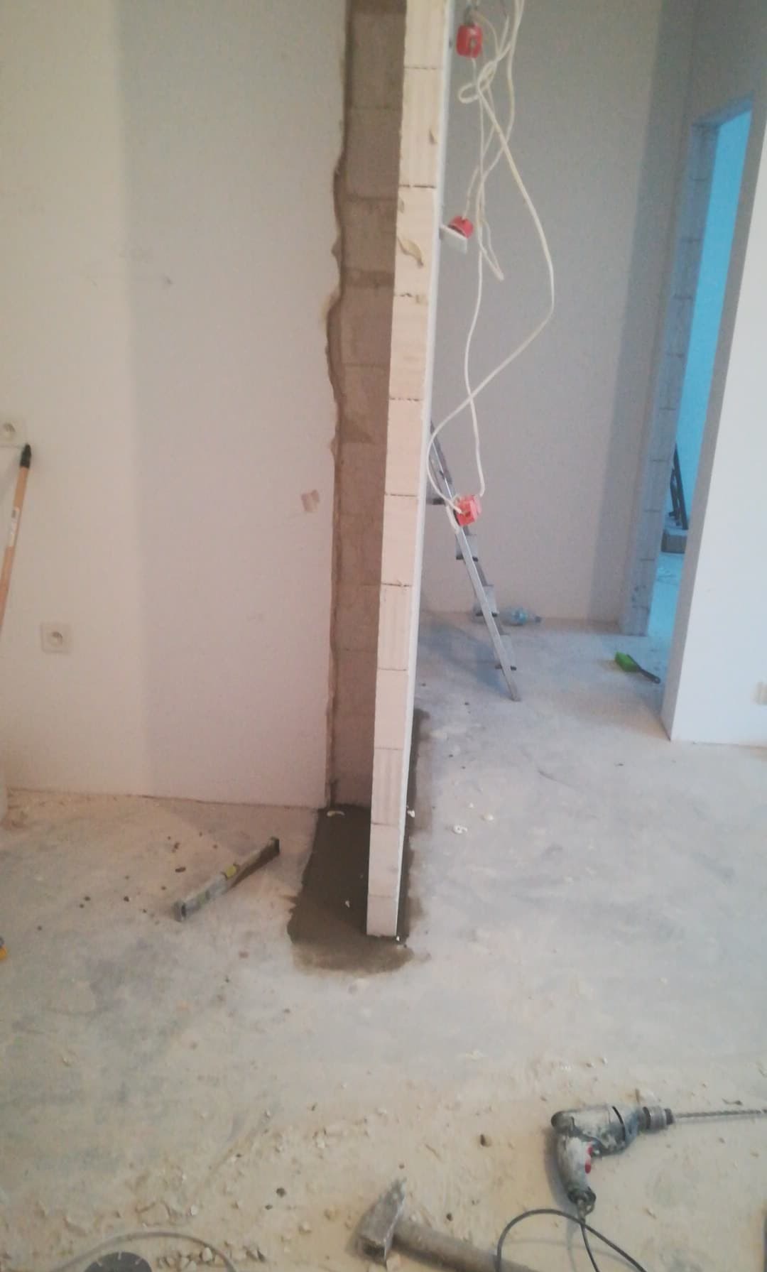 Wznoszenie ścianki działowej z bloczków, widoczne narzędzia, poziomica, wiertarka, młotek, przewody elektryczne wystające ze ściany, drabina w tle.