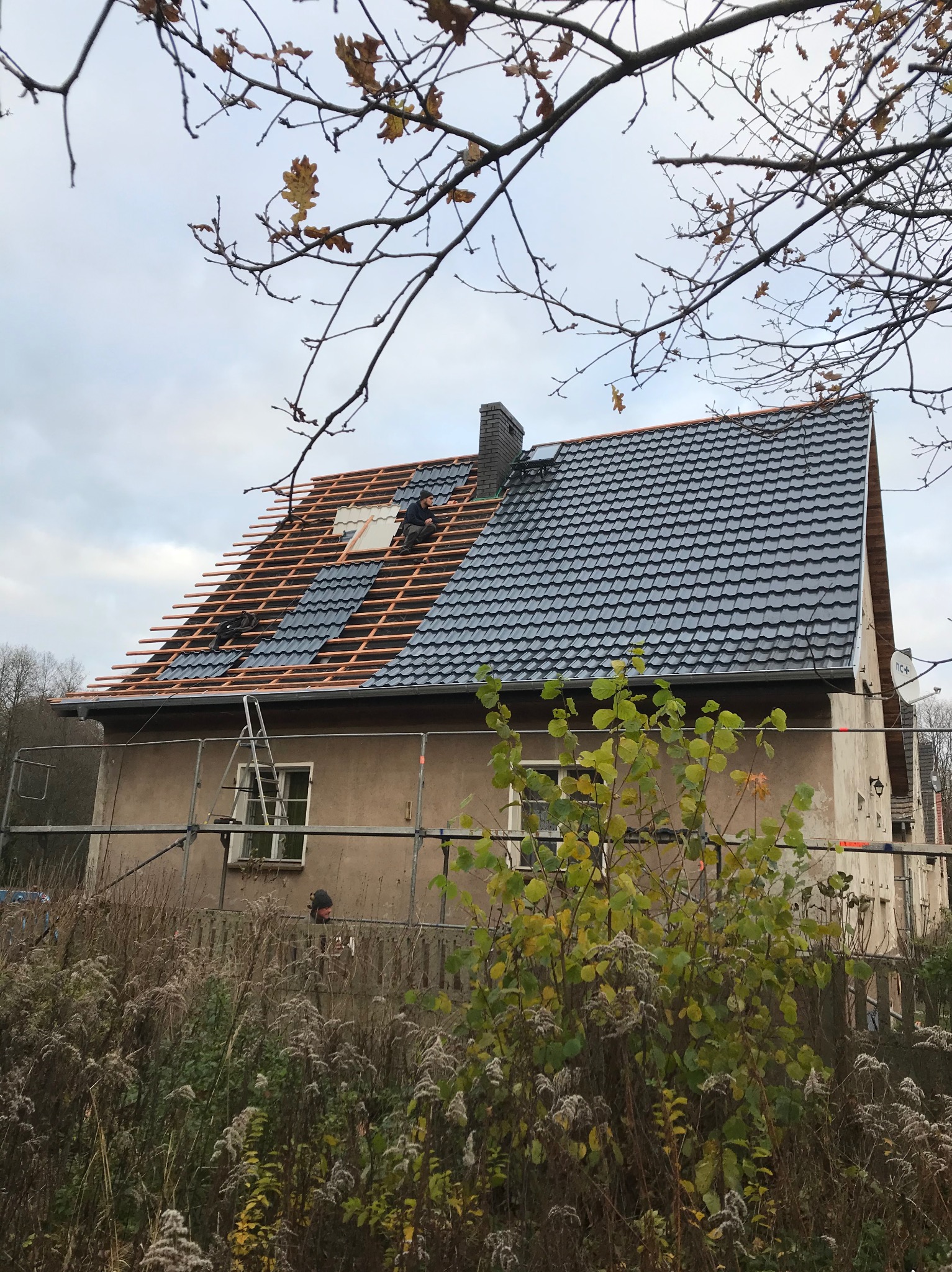 Dom jednorodzinny w trakcie krycia dachu blachodachówką modułową; widoczne łaty, częściowo ułożone panele, komin z cegły, rusztowanie i pracujący dekarz.