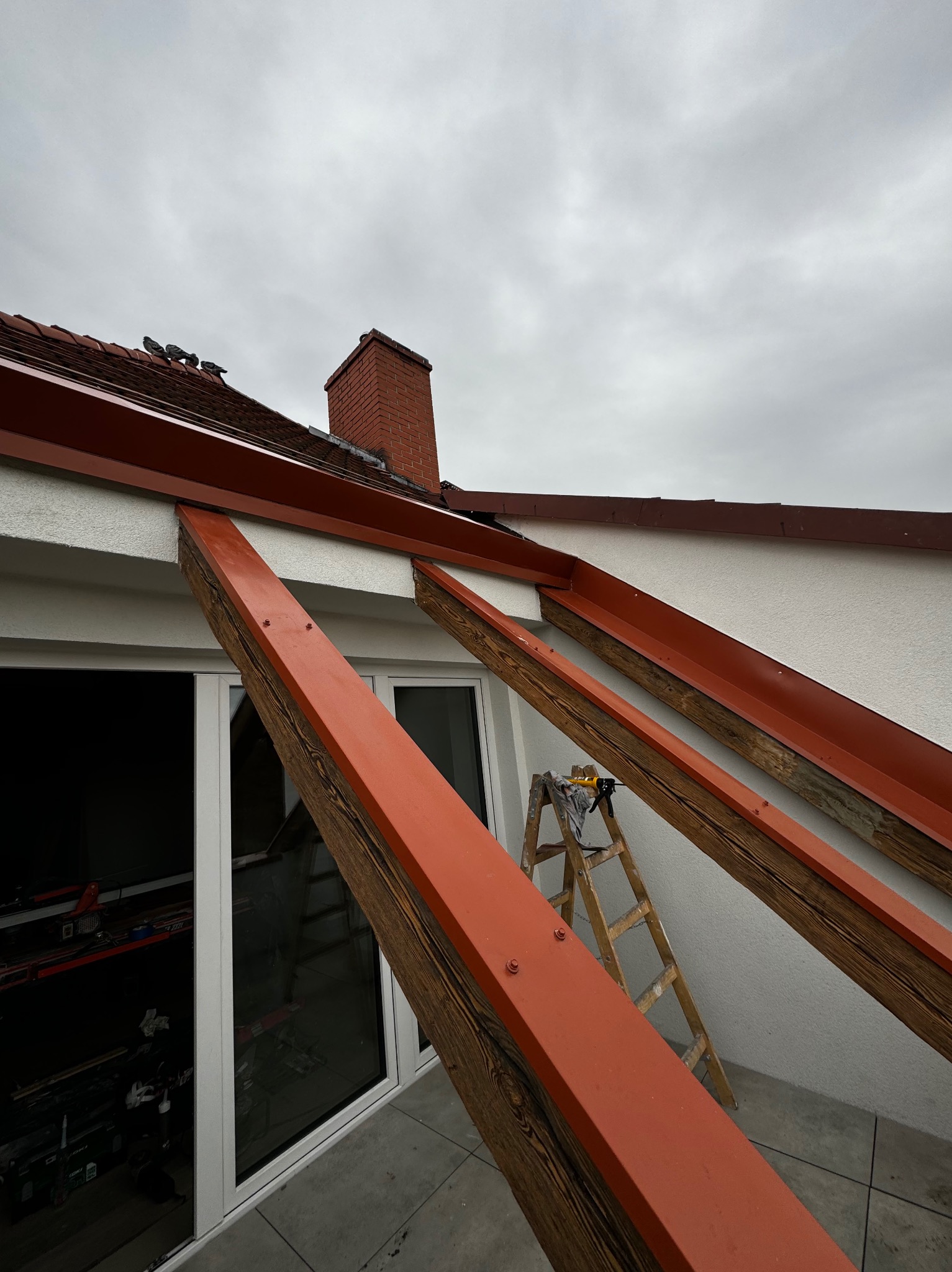 Otwarcie dachu z zachowaniem drewnianej więźby, widoczne elementy konstrukcyjne pomalowane na czerwono, drabina oparta o ścianę, dachówka i komin w tle.