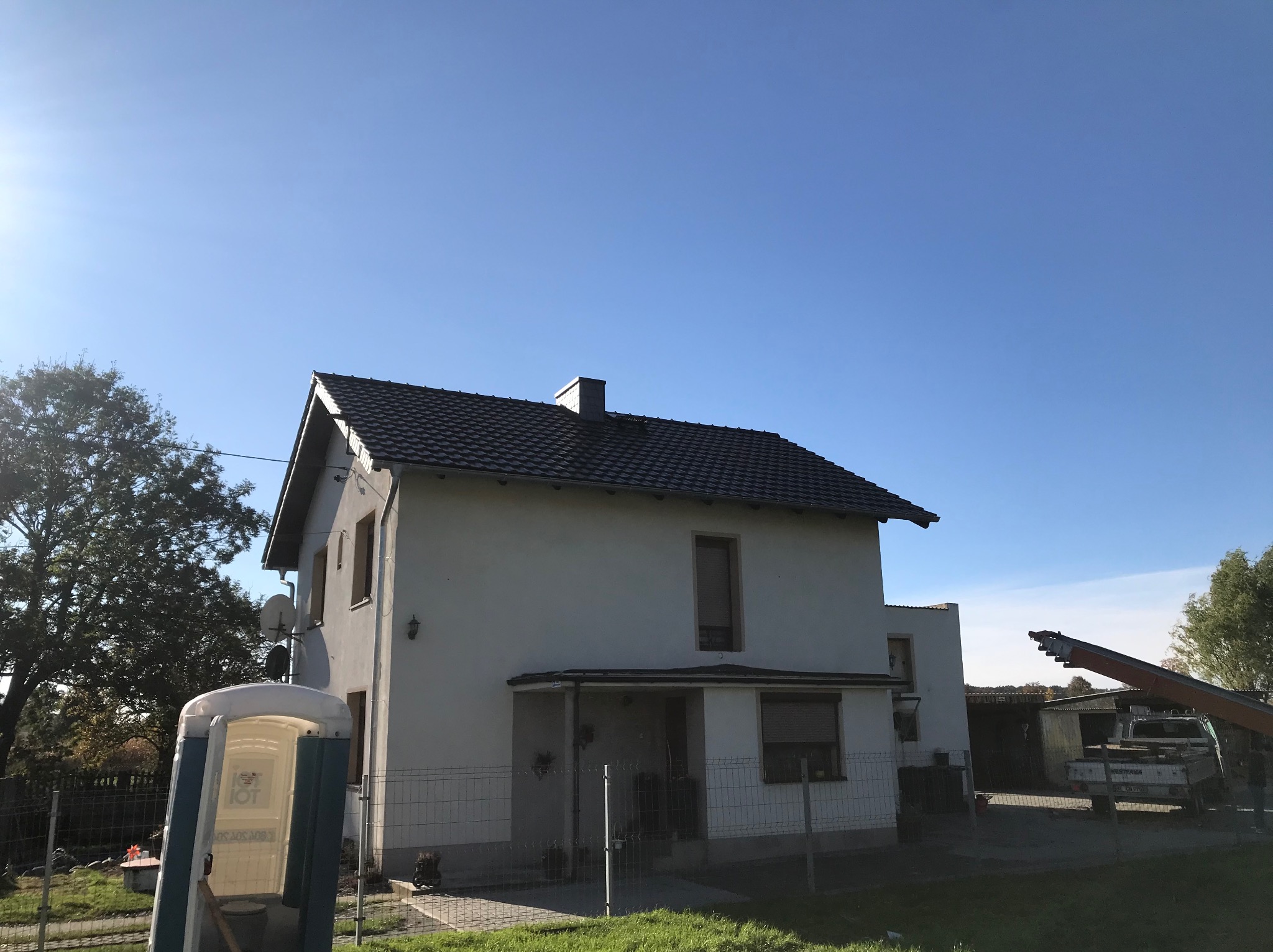 Dom jednorodzinny z nowym, ciemnym dachem z dachówki, widoczny podnośnik koszowy i przenośna toaleta na posesji.