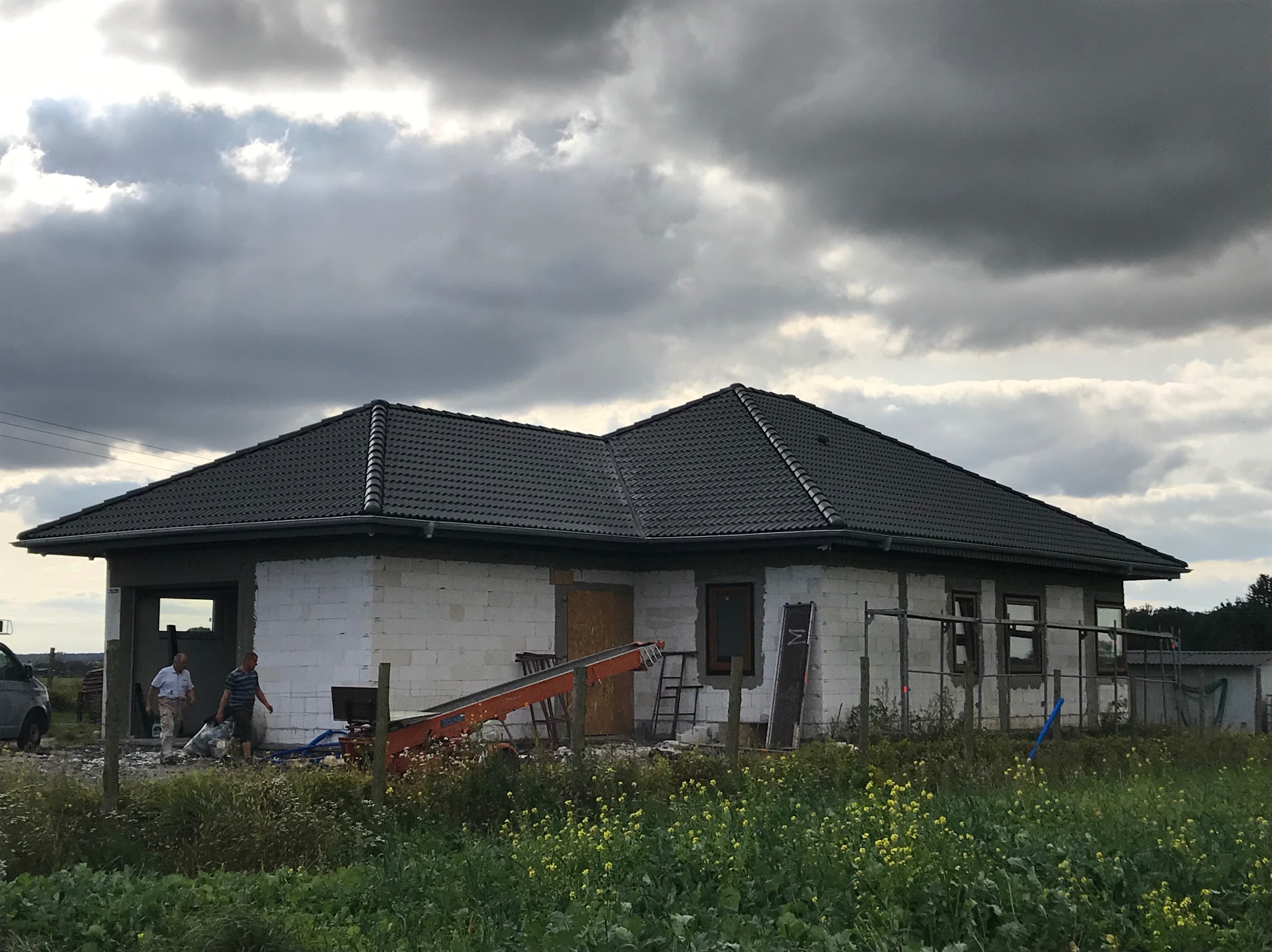 Dom jednorodzinny w trakcie budowy z nowym, grafitowym dachem z dachówki ceramicznej; widoczni dwaj mężczyźni w pobliżu wejścia, pomarańczowa taśmociągowa winda dekarska i pochmurne niebo w tle.