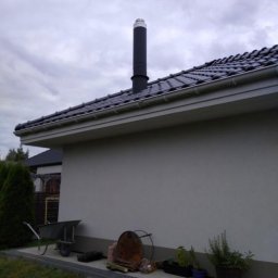 Frez-Tom - Czarny komin z białym zakończeniem wystający z ciemnej, ceramicznej dachówki nowego budynku, z taczką i różnymi przedmiotami leżącymi na ziemi wzdłuż ściany.