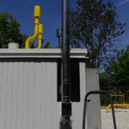 Zewnętrzny, stalowy przewód kominowy zamontowany na ścianie budynku z widoczną instalacją gazową w kolorze żółtym i drabiną opartą o ścianę. W tle drzewa i błękitne niebo.