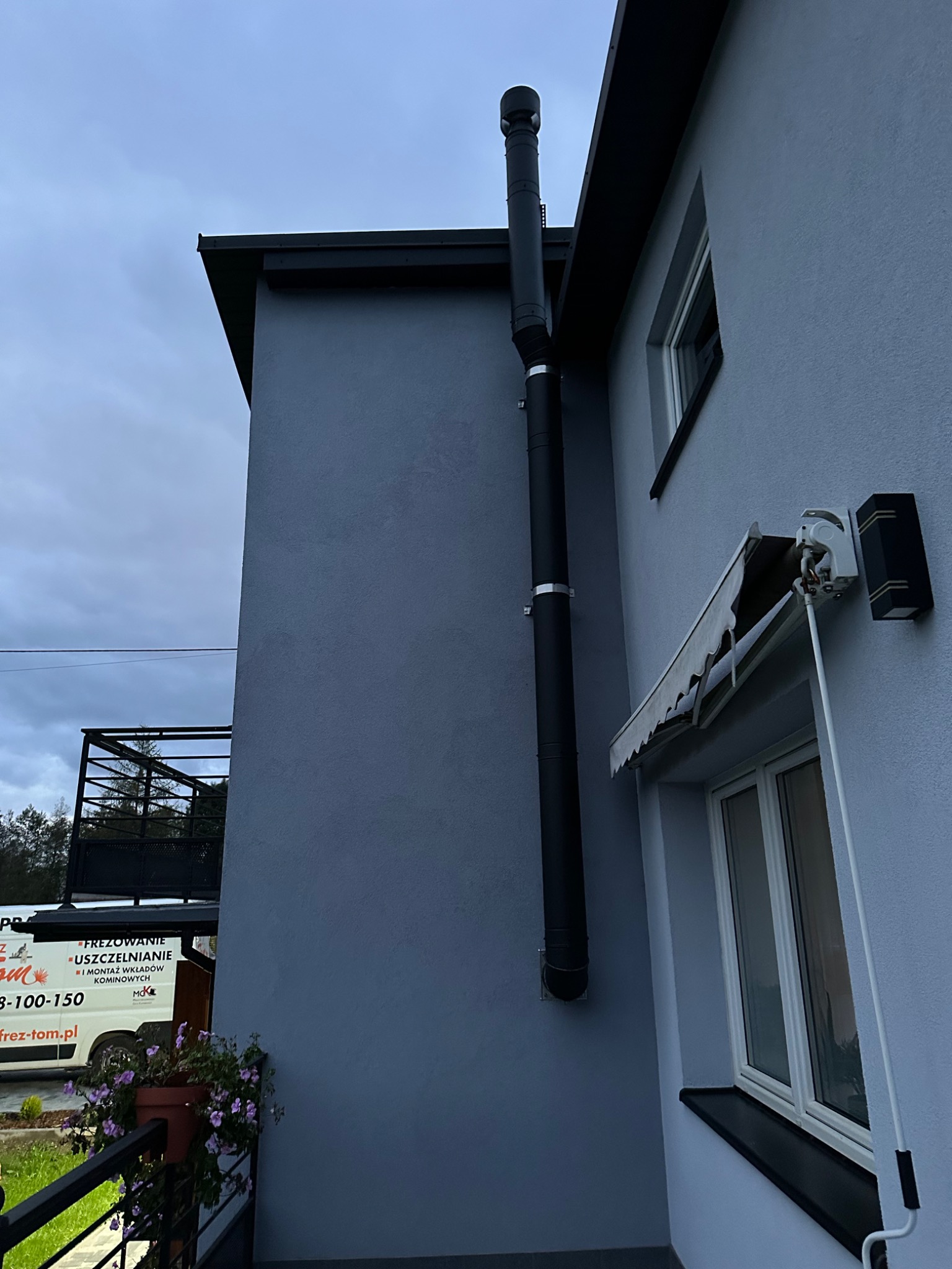 Zewnętrzny, czarny system odprowadzania spalin zamontowany na ścianie domu o szarej elewacji, widoczny z boku, z fragmentem okna i markizy.