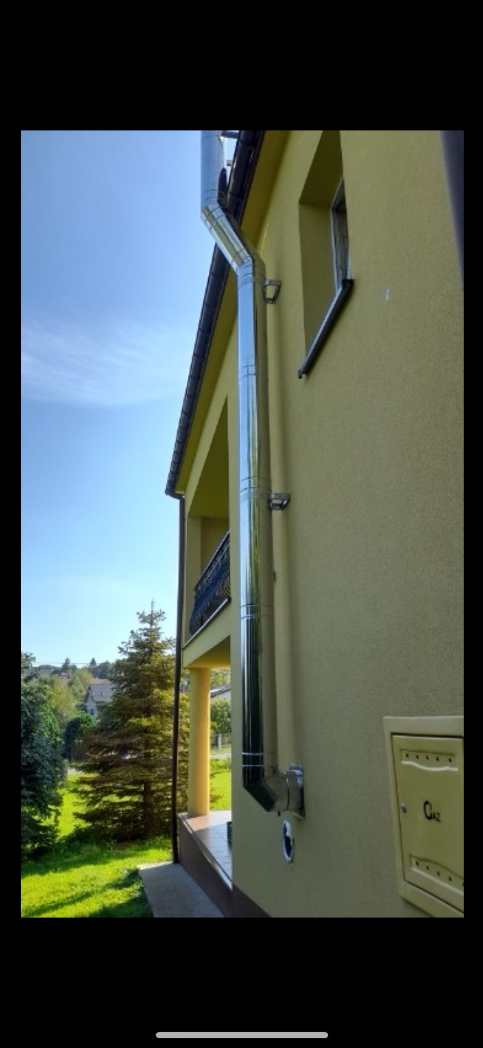 Błyszczący, metalowy przewód kominowy zamontowany na zewnętrznej ścianie budynku z żółtą elewacją, widok z dołu w słoneczny dzień.