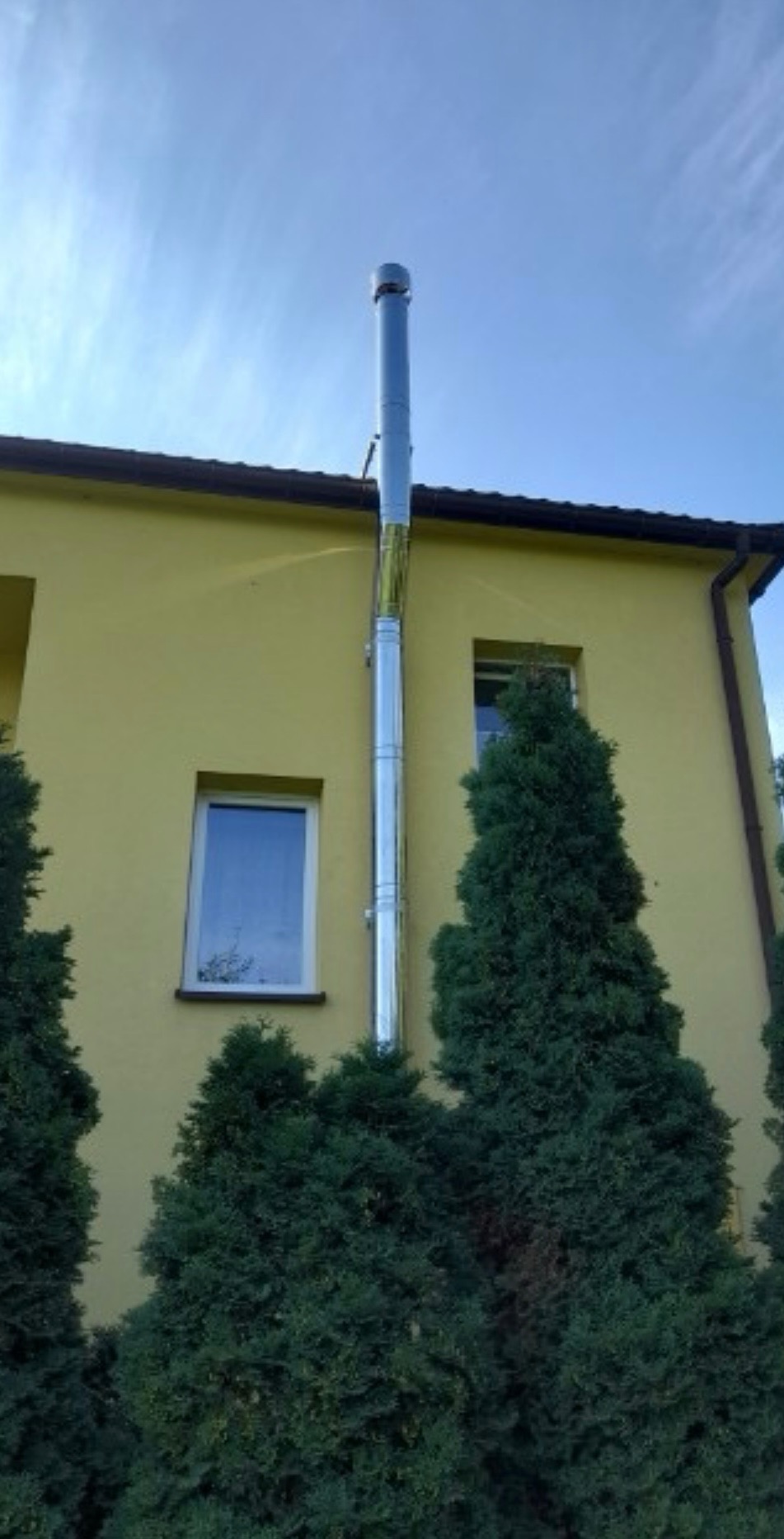 Zewnętrzny, metalowy przewód spalinowy zamontowany na żółtej ścianie budynku, widoczny z poziomu gruntu, z tłem błękitnego nieba i częściowo zasłonięty przez zielone krzewy.