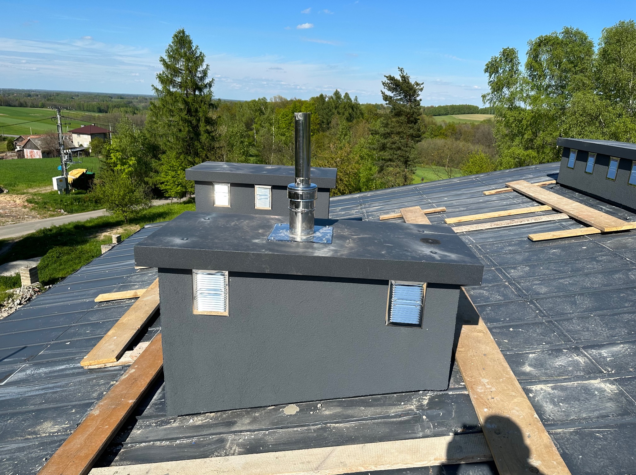 Szare kominy wentylacyjne z metalowymi wylotami na dachu z blachy, drewniane deski zabezpieczające, widok na zielone drzewa i krajobraz w tle.