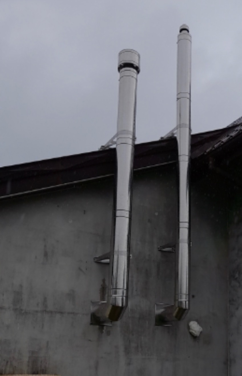 Dwa stalowe przewody odprowadzające spaliny zamocowane do ściany budynku, widoczne mocowania i zakończenia przewodów, pochmurne niebo w tle.