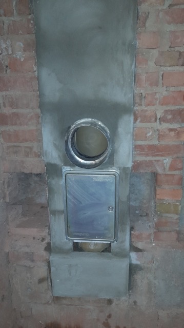 Murowany przewód z otworem wentylacyjnym i metalową klapką rewizyjną, otoczony świeżym tynkiem na tle ceglanej ściany.