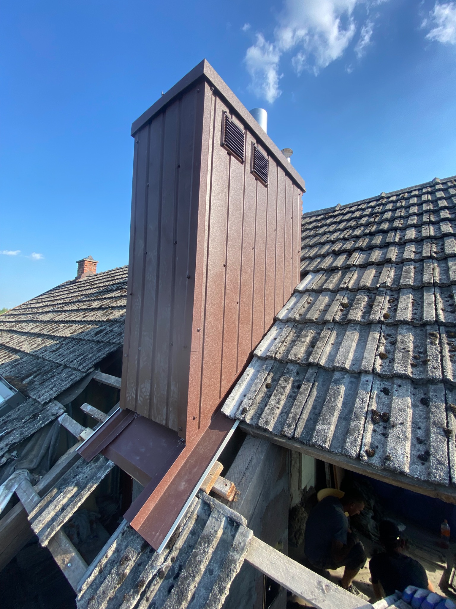 Montaż nowego, brązowego komina z blachy na dachu pokrytym eternitem; widoczne otwory wentylacyjne w kominie i fragment nieba.