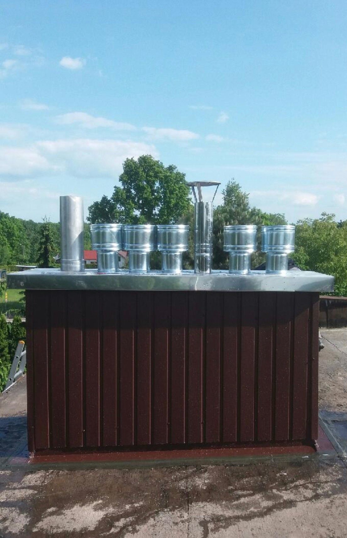 Brązowy komin z siedmioma metalowymi wylotami o różnej konstrukcji na tle błękitnego nieba i zieleni drzew.