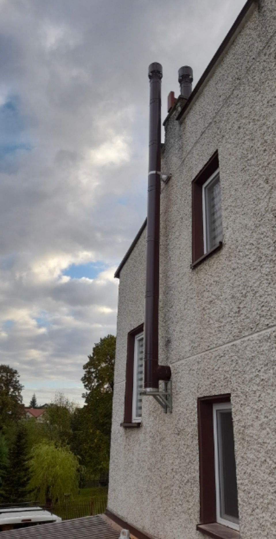 Zewnętrzny, brązowy system odprowadzania spalin zamontowany na elewacji budynku z oknami o brązowych ramach, widok z dołu na tle pochmurnego nieba.