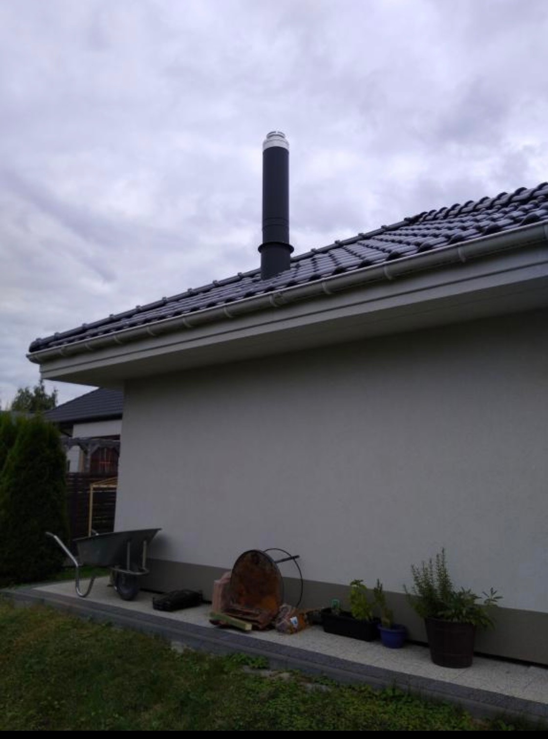 Czarny komin z białym zakończeniem wystający z ciemnej, ceramicznej dachówki nowego budynku, z taczką i różnymi przedmiotami leżącymi na ziemi wzdłuż ściany.