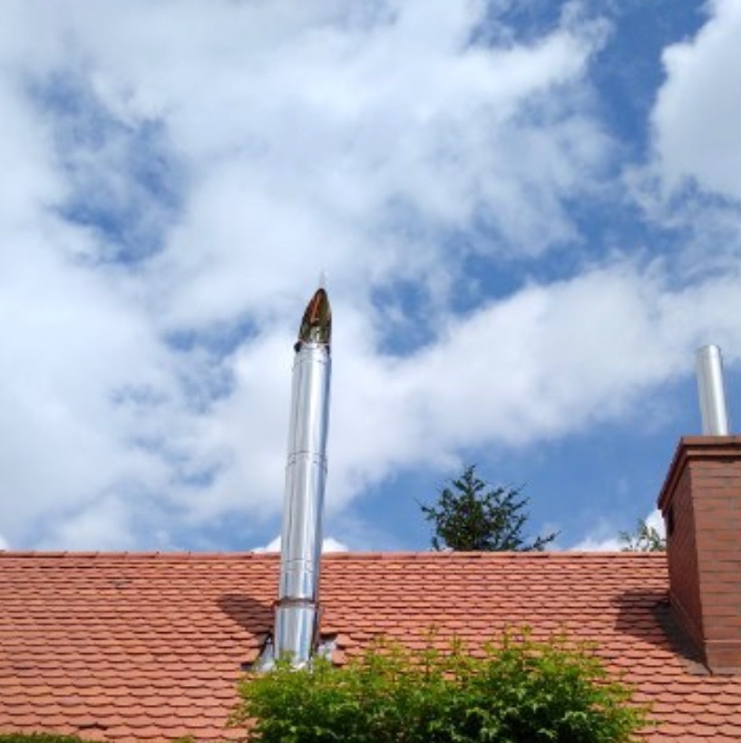 Błyszczący, metalowy przewód kominowy wystający ponad dach z czerwonej dachówki, na tle błękitnego nieba z białymi chmurami. Widoczny fragment tradycyjnego komina z cegły.