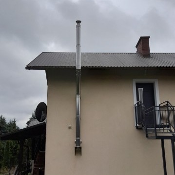 Zewnętrzny, stalowy system odprowadzania spalin zamontowany przy ścianie budynku z brązowym dachem i schodami wejściowymi. Widoczne elementy montażowe oraz fragment komina na dachu.