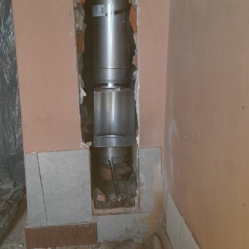 Montaż stalowego wkładu kominowego wewnątrz murowanego komina. Widoczne uszkodzenia ściany wokół otworu kominowego i gruz na dole.