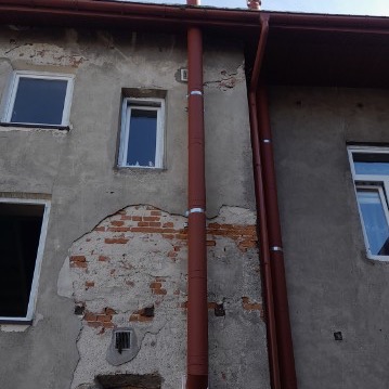 Uszkodzona elewacja budynku z widoczną cegłą, pionowe rury spustowe w kolorze ceglastym przymocowane do ściany za pomocą metalowych obejm, dwa okna z białymi ramami, fragment dachu z rynną w górnej...