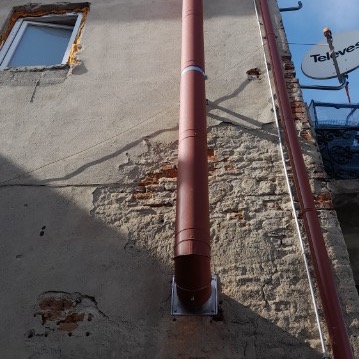 Stara, otynkowana ściana budynku z widocznymi fragmentami cegieł, z zamontowaną rurą spustową w kolorze brązowym, przytwierdzoną metalowym uchwytem. W tle widoczny fragment okna i antena satelitarna.