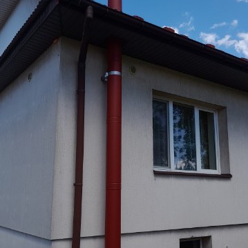 Czerwony, metalowy przewód kominowy na zewnątrz jasnego budynku z oknem, zamocowany za pomocą srebrnej obejmy, widoczny fragment dachu i niebieskie niebo z chmurami.