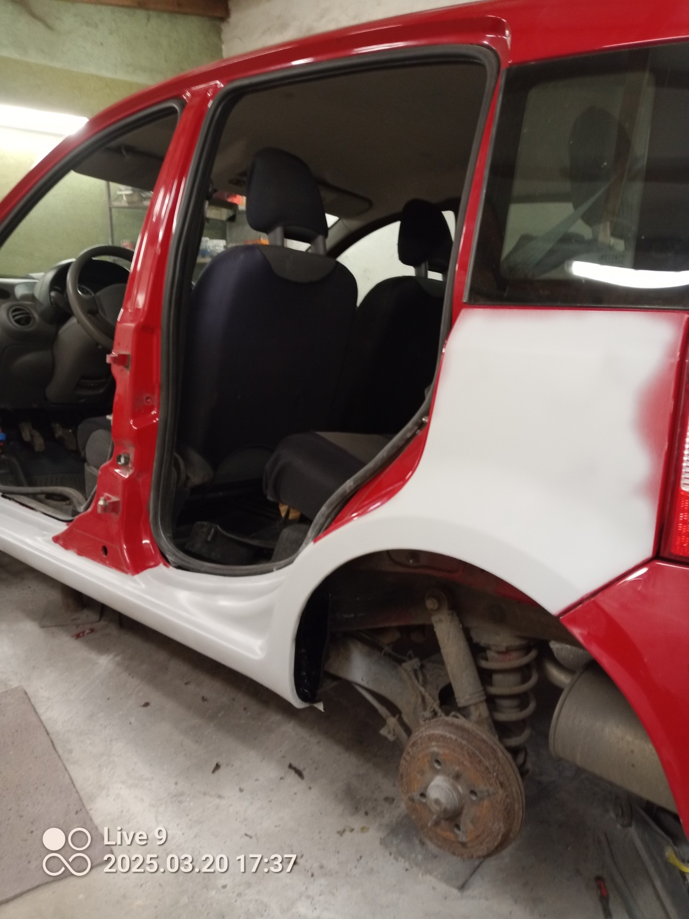 Czerwony samochód w trakcie naprawy blacharskiej, z elementami w szarym podkładzie, brak koła, widoczne zawieszenie. Wnętrze pojazdu z widocznymi fotelami.