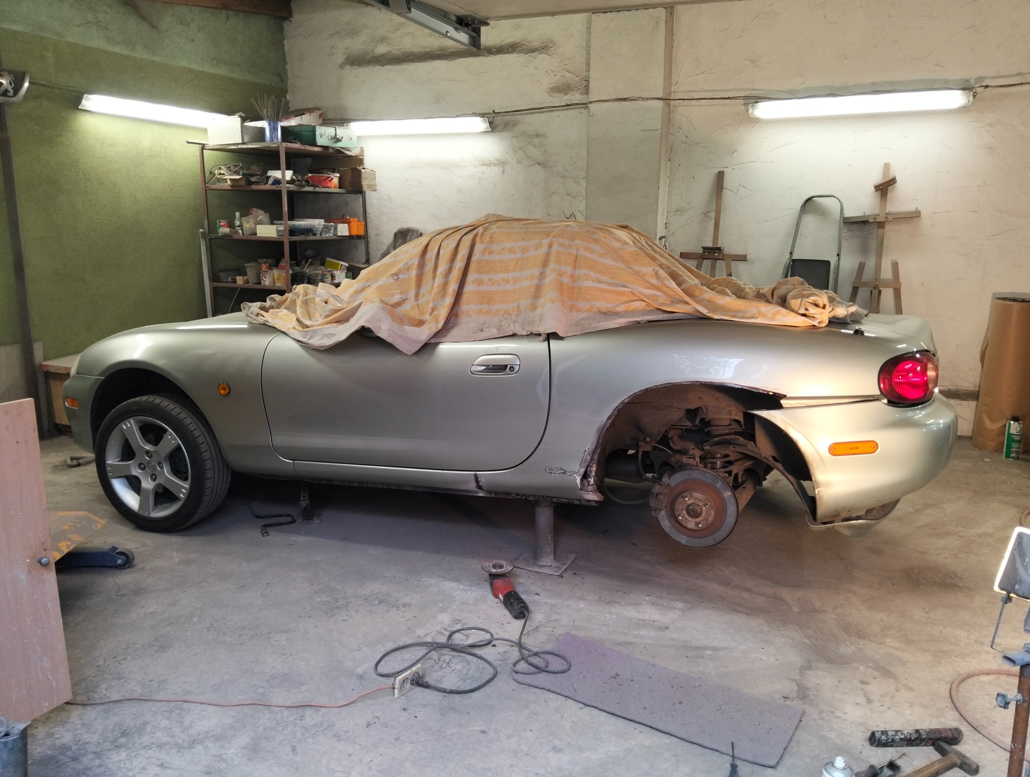 Srebrny samochód na podnośniku w warsztacie, z odsłoniętym nadkolem i narzędziami obok. Auto przykryte materiałem, sugerujące prace blacharskie lub lakiernicze.