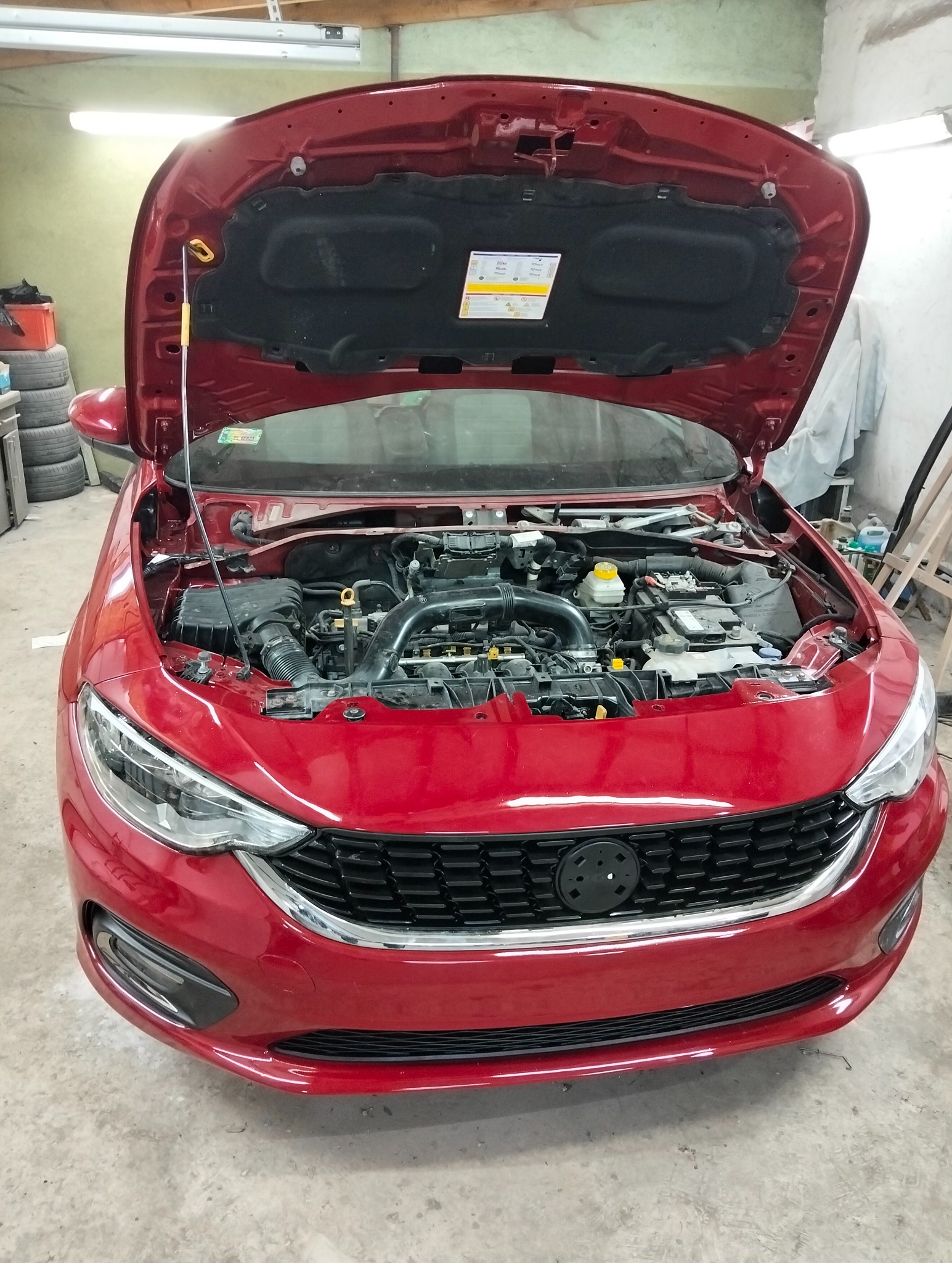 Czerwony samochód z otwartą maską w warsztacie. Widoczny silnik i elementy mechaniczne. Detal: żółty uchwyt podpory maski.