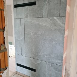 BB-PROBUD - Ściana wykończona płytkami imitującymi kamień w odcieniach szarości z czarnymi wnękami wentylacyjnymi, widoczne surowe wykończenie ścian bocznych z cegły i płyty gipsowej.