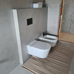 BB-PROBUD - Nowoczesna łazienka w trakcie wykańczania z szarą płytką, wiszącą toaletą i bidetem z czarną armaturą, zabezpieczona podłoga kartonem falistym.