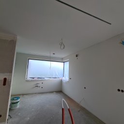 BB-PROBUD - Surowe wnętrze w trakcie prac wykończeniowych: ściany częściowo pokryte płytami gipsowo-kartonowymi, widoczne puszki elektryczne, okno zabezpieczone folią, na podłodze wiadro z farbą i rury...